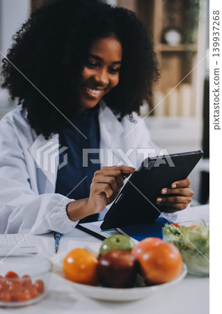 Young nutritionist showing bell peppers during an online video call consulting about healthy eating, sitting at home at a table with vegetables, laptop and measuring tape Young nutritionist showing bell peppers during an online video call consulting about healthy eating, sitting at home at a table with vegetables, laptop and measuring tape 139937268