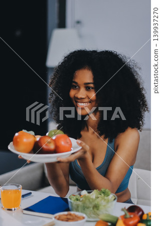 Young nutritionist showing bell peppers during an online video call consulting about healthy eating, sitting at home at a table with vegetables, laptop and measuring tape 139937270