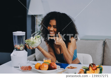 Young nutritionist showing bell peppers during an online video call consulting about healthy eating, sitting at home at a table with vegetables, laptop and measuring tape 139937271