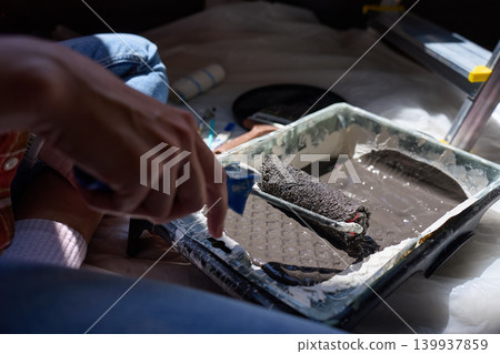 Woman dips paint roller in tray with dark paint on protective plastic sheet 139937859