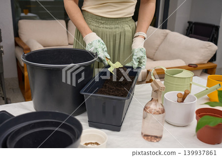 Woman prepares planter with potting soil using hand trowel at garden table 139937861