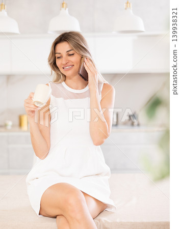 Delightful blonde drinking coffee and talking on the phone while sitting on kitchen table Delightful blonde drinking coffee and talking on the phone while sitting on kitchen table 139938321