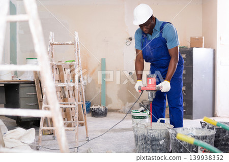 Construction worker mixing dry mortar with handheld mixer in bucket in overhauls 139938352