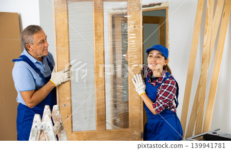 Two contractor repairmen in a blue uniform holding a door in polyethylene before installation in the house 139941781