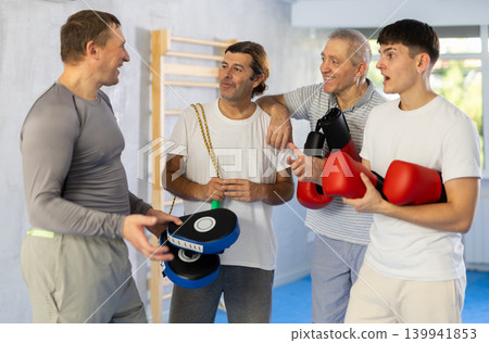 Four men of different ages in gym at group training session with sports equipment speaking Four men of different ages in gym at group training session with sports equipment speaking 139941853
