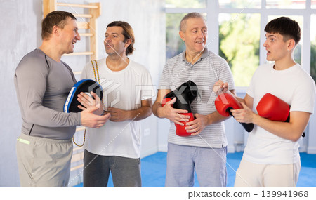 Four men of different ages in gym at group training session with sports equipment speaking Four men of different ages in gym at group training session with sports equipment speaking 139941968