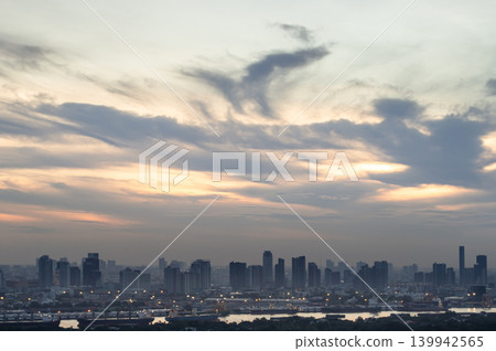 Sweeping clouds part to reveal an amber horizon shifting from dusk to white. Below, skyscrapers cut a sharp silhouette as city lights flicker. Along the dark riverfront, cargo ships rest quietly in Sweeping clouds part to reveal an amber horizon shifting from dusk to white. Below, skyscrapers cut a sharp silhouette as city lights flicker. Along the dark riverfront, cargo ships rest quietly in 139942565