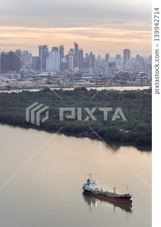 A solitary cargo vessel glides across the dawn-lit Chao Phraya River. In the foreground, the lush Bang Kachao peninsula forms a dense natural sanctuary. Beyond the greenery, the sprawling Bangkok A solitary cargo vessel glides across the dawn-lit Chao Phraya River. In the foreground, the lush Bang Kachao peninsula forms a dense natural sanctuary. Beyond the greenery, the sprawling Bangkok 139942714