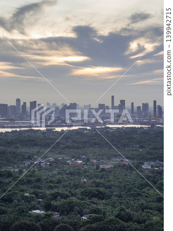 A lush, emerald forest dominates the foreground, creating a natural oasis. Beyond the trees, a silver river catches the pale dawn light. On the opposite bank, a towering cityscape rises through the A lush, emerald forest dominates the foreground, creating a natural oasis. Beyond the trees, a silver river catches the pale dawn light. On the opposite bank, a towering cityscape rises through the 139942715