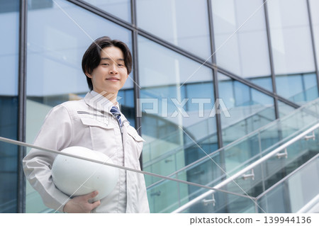 A young male engineer smiles against a backdrop of city buildings, representing a construction career image: job hunting, career, growth, and challenges. A young male engineer smiles against a backdrop of city buildings, representing a construction career image: job hunting, career, growth, and challenges. 139944136