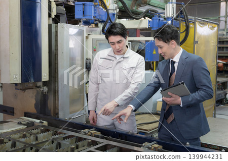 In a manufacturing plant, technicians in work clothes and sales staff are looking at the machinery and discussing estimates, repair costs, and maintenance fees. 139944231