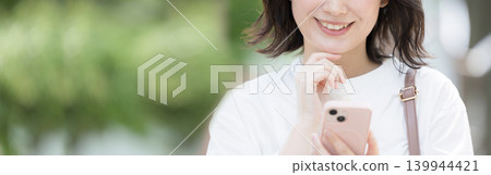 一張生活方式照片，照片中一位面帶微笑的女性手持智慧型手機，置身於綠意盎然的戶外，但表情卻有些困惑、迷茫或擔憂，似乎正在查看地圖等等。 139944421