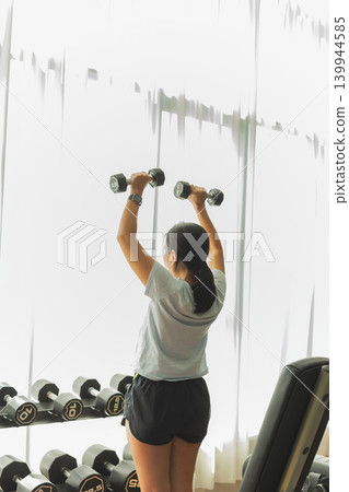 Rear view of woman performing an overhead dumbbell press exercise at the gym. Rear view of woman performing an overhead dumbbell press exercise at the gym. 139944585