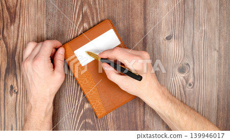 hand writing with a pen on a white rectangle of a closed brown notebook hand writing with a pen on a white rectangle of a closed brown notebook 139946017