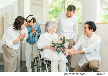 A three-generation family visits their grandmother in a care facility and presents her with a bouquet of flowers as a get-well gift. A three-generation family visits their grandmother in a care facility and presents her with a bouquet of flowers as a get-well gift. 139946604