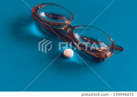 White pill under the magnifying lens of reading glasses lying on a blue background, illustrating the importance of careful study of medication instructions and transparency in healthcare. White pill under the magnifying lens of reading glasses lying on a blue background, illustrating the importance of careful study of medication instructions and transparency in healthcare. 139946855
