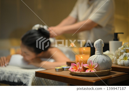Focus on spa equipment and relaxing candlelight woman receiving traditional herbal compress massage in a serene spa Focus on spa equipment and relaxing candlelight woman receiving traditional herbal compress massage in a serene spa 139947187