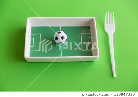 Small soccer ball on football field plate with fork on bright green sports nutrition concept. Small soccer ball on football field plate with fork on bright green sports nutrition concept. 139947339