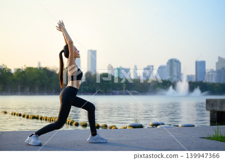 Woman raises arms stretching by lake with city view at sunrise 139947366