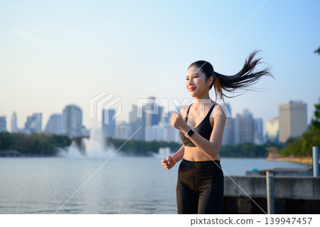 Fit woman jogging by lake with city skyline at sunrise Fit woman jogging by lake with city skyline at sunrise 139947457
