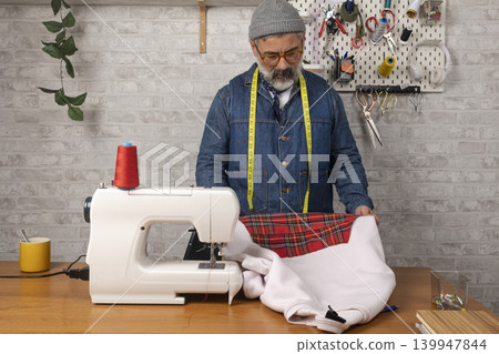 Adult man sewing plaid fabric on a sewing machine Adult man sewing plaid fabric on a sewing machine 139947844