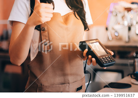 Friendly Asian female barista receives a credit card from a customer for payment. Both women are smiling in a warmly lit coffee shop with a rustic interior. Cashless payments. Small family business Friendly Asian female barista receives a credit card from a customer for payment. Both women are smiling in a warmly lit coffee shop with a rustic interior. Cashless payments. Small family business 139951637