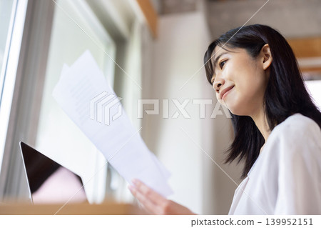 A young Japanese woman working on her computer with documents in hand by a bright window. A young Japanese woman working on her computer with documents in hand by a bright window. 139952151