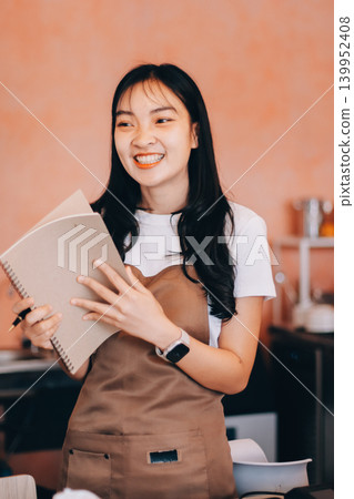 Friendly Asian female barista receives a credit card from a customer for payment. Both women are smiling in a warmly lit coffee shop with a rustic interior. Cashless payments. Small family business Friendly Asian female barista receives a credit card from a customer for payment. Both women are smiling in a warmly lit coffee shop with a rustic interior. Cashless payments. Small family business 139952408