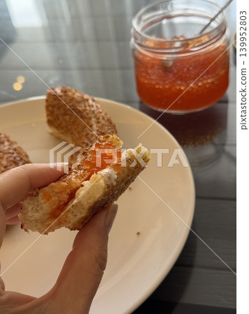 Red caviar spread on sesame bagel halves with jar on table, close-up of person holding piece Red caviar spread on sesame bagel halves with jar on table, close-up of person holding piece 139952803