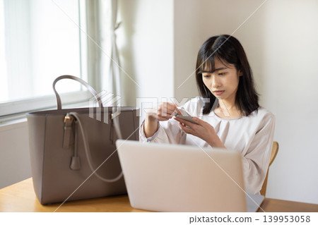 A young Japanese woman places her laptop and bag on a table and checks her smartphone. A young Japanese woman places her laptop and bag on a table and checks her smartphone. 139953058