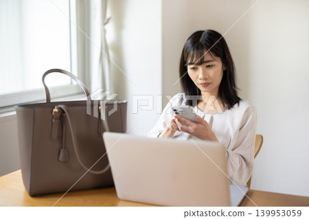 A young Japanese woman places her laptop and bag on a table and checks her smartphone. 139953059