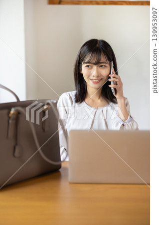 A young Japanese woman places her laptop and bag on a table and talks on her smartphone. 139953097