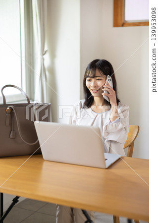 A young Japanese woman places her laptop and bag on a table and talks on her smartphone. A young Japanese woman places her laptop and bag on a table and talks on her smartphone. 139953098