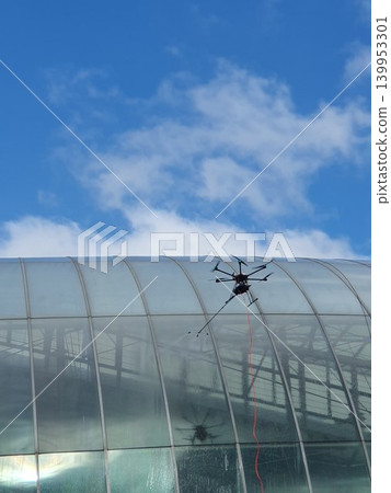 A tethered inspection drone hovers beside a curved glass roof of a modern building, conducting maintenance and window cleaning under a bright blue sky with scattered clouds, urban drone technology. 139953301