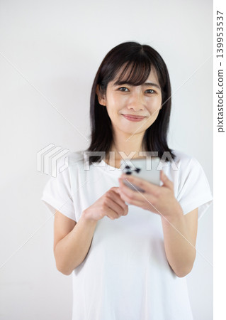 一位年輕的日本女子在白色背景前手持智慧型手機微笑。 139953537