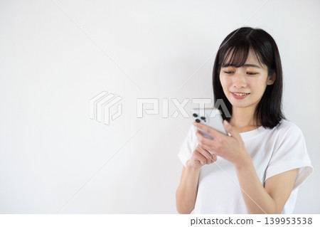 一位年輕的日本女子在白色背景前手持智慧型手機微笑。 139953538