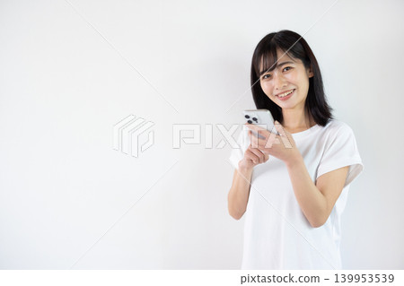 一位年輕的日本女子在白色背景前手持智慧型手機微笑。 139953539
