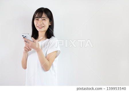 一位年輕的日本女子在白色背景前手持智慧型手機微笑。 139953540