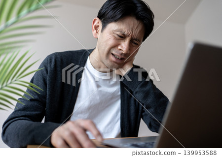 Japanese man operating a computer while experiencing neck pain and cervical spine discomfort. Japanese man operating a computer while experiencing neck pain and cervical spine discomfort. 139953588