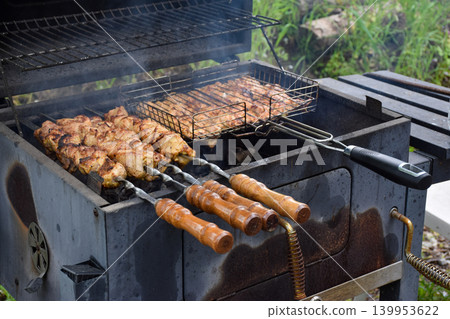 Grilled meat skewers on a barbecue grill. Smoke rises from the cooking food. Juicy pieces of meat are arranged in a metal grilling basket. 139953622