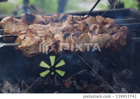 Grilled meat skewers on a barbecue grill. Smoke rises from the cooking food. Juicy pieces of meat are arranged in a metal grilling basket. Grilled meat skewers on a barbecue grill. Smoke rises from the cooking food. Juicy pieces of meat are arranged in a metal grilling basket. 139953741