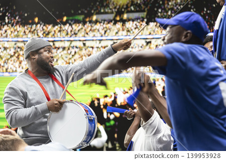Dedicated football fan leader directing blue supporters with drum and megaphone in stadium terrace during loud coordinated soccer chant. Organized supporters celebrate team spirit through cheering. Dedicated football fan leader directing blue supporters with drum and megaphone in stadium terrace during loud coordinated soccer chant. Organized supporters celebrate team spirit through cheering. 139953928
