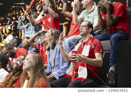 Red team soccer fans reacting to close call in stadium stands, hands on head, scarves and anxious crowd during live football match. Anxious supporters show tension and suspense, inclusive fans. Red team soccer fans reacting to close call in stadium stands, hands on head, scarves and anxious crowd during live football match. Anxious supporters show tension and suspense, inclusive fans. 139953931