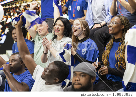 Blue team football supporters clapping and raising arms in crowded stadium stands during exciting live soccer match. Sports fans with face paint creating loud crowd chants, game excitement. 139953932