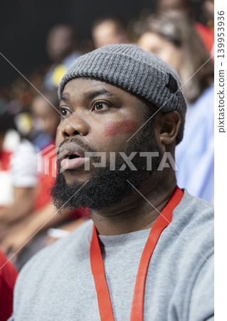 Close portrait of worried Black football fan in gray beanie watching tense live soccer match from stadium stands with face paint, anticipation to score goal. Crowded stadium in total silence. Close portrait of worried Black football fan in gray beanie watching tense live soccer match from stadium stands with face paint, anticipation to score goal. Crowded stadium in total silence. 139953936