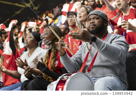 Frustrated football supporter with drum reacting with open hands in red stadium crowd during dramatic live soccer match moment. Black drummer experiences silence and intense game pressure. Frustrated football supporter with drum reacting with open hands in red stadium crowd during dramatic live soccer match moment. Black drummer experiences silence and intense game pressure. 139953942