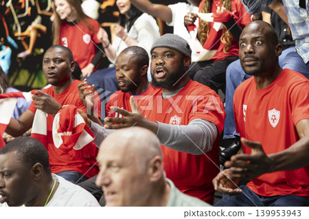 Diverse sports fans reacting with suspense during live football match in stadium stands, with red shirts and focused expressions. Anxious supporters watching decisive final minutes in silence. Diverse sports fans reacting with suspense during live football match in stadium stands, with red shirts and focused expressions. Anxious supporters watching decisive final minutes in silence. 139953943