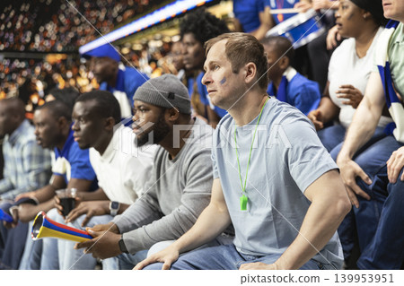 Tense soccer fans watching decisive moment during live football match in stadium filled with suspense and anticipation. Worried fans holding breath during crucial penalty kick, serious crowd. 139953951