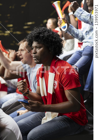 Vertical portrait of football supporter checking smartphone during tense live soccer match in crowded stadium stands. Fan with wig uses betting app for in-play risky wager, live betting. Vertical portrait of football supporter checking smartphone during tense live soccer match in crowded stadium stands. Fan with wig uses betting app for in-play risky wager, live betting. 139953971