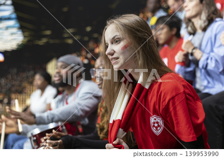 Focused young female football fan in red shirt watching tense live soccer match from stadium stands with scarf and crowd behind. Supporters of the red team show intense pressure, final minutes. 139953990